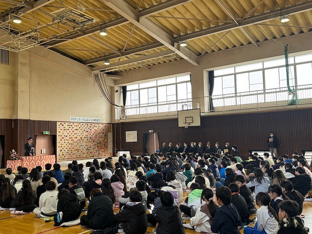 体育館で、多くの生徒が床に座って記念式典に参加し、横には数人の来賓の方々が座り、壇上でスーツ姿の市長が話しているのを聞いている様子の写真