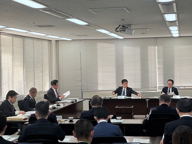 白いブラインドがかかった部屋で、複数の男性が長机を囲んで座っており、中央の二人の男性が書類を確認している会議風景の写真