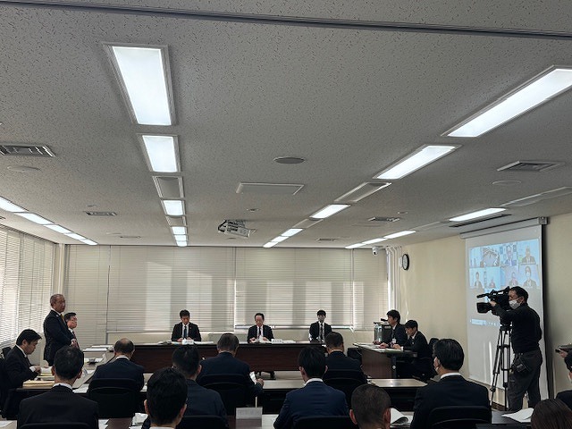 大会議室で多くのスーツ姿の男女がコの字型のテーブルに座っており、その様子を撮影するカメラマンが写っている会議風景の写真