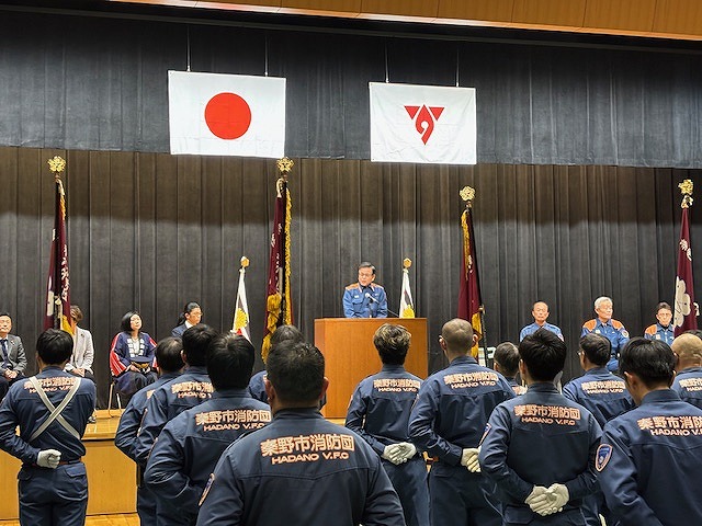 日本の国旗と市章の旗が掲げられた壇上で、制服を着た市長が演台に立ち、整列した秦野市消防団の団員たちに向かって話している式典の写真
