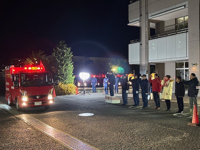 消防車両と複数の人が夜の屋外に並んでおり、全員が敬礼をしている消防団歳末火災特別警戒激励式の様子の写真