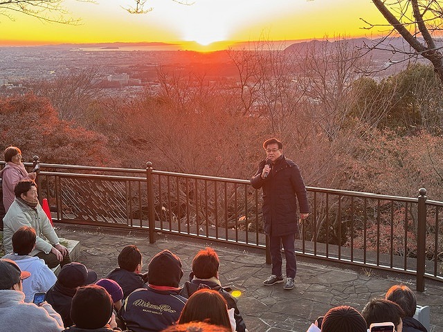 日が昇り始めた早朝、山頂の展望台で黒い防寒着を着た市長がマイクを持って挨拶をしており、集まった参加者が日の出の美しい景色を眺めながら話を聞いている新年の幕開けを捉えた写真
