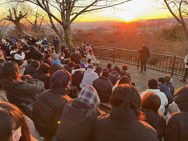 ニューイヤーウォークに参加する大勢の人々が、山頂の展望台のような場所に集まって、初日の出を背景に市長がマイクで話している様子を捉えた写真