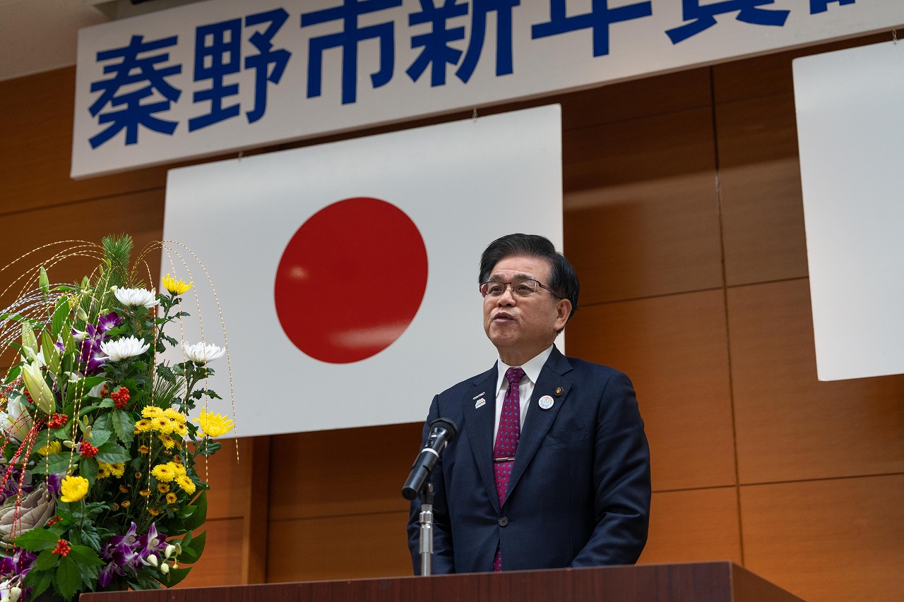 「秦野市新年賀詞交歓会」と書かれた横断幕と国旗が飾られた式典の壇上で、スーツ姿の男市長がマイクに向かって挨拶を行っている様子をアップで撮影した写真