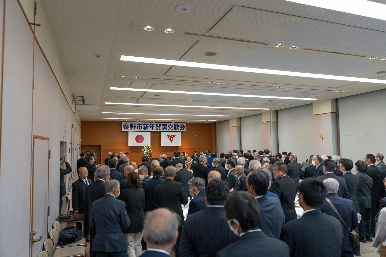 室内のステージ上に「秦野市新年賀詞交歓会」と書かれた横断幕が掲げられており、多くの参加者が集まって立っている会場の様子を後方から撮影した写真