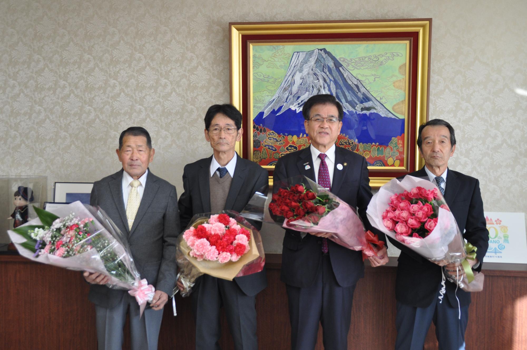 金色の額縁に入った山頂に雪が積もった大きな山の絵画が壁に飾られた落ち着いた背景の市役所の応接室で、スーツ姿の市長を中央に3人の男性が花束を持って並び、カメラに向かって記念撮影に応じている写真