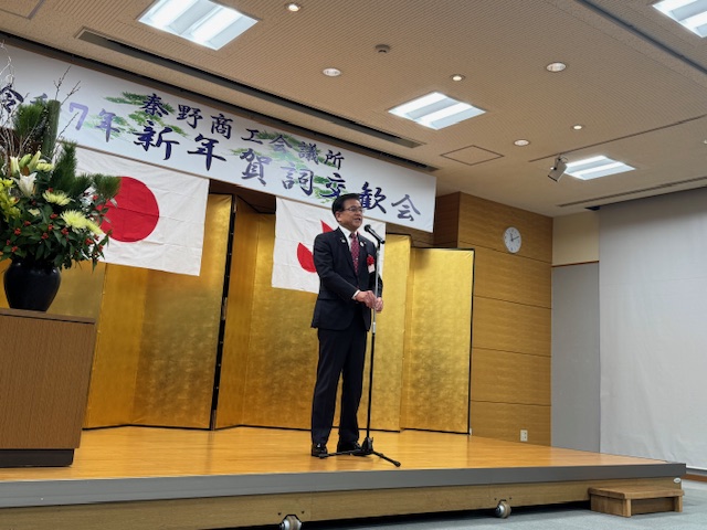 「秦野市商工会議所令和7年新年賀詞交歓会」と書かれた横断幕と2枚の旗が飾られ、金屏風が設置された小さな舞台上で、スーツ姿の市長がマイクスタンドの前に立ち挨拶をしている様子の写真