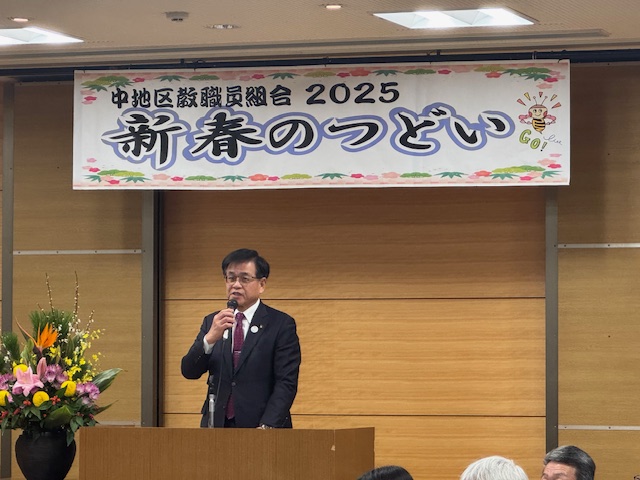 「中地区教職員組合2025新春のつどい」と書かれた横断幕が掲げられている会場の演壇に立つスーツ姿の市長が、右手にマイクを持って挨拶をしている様子を写した写真