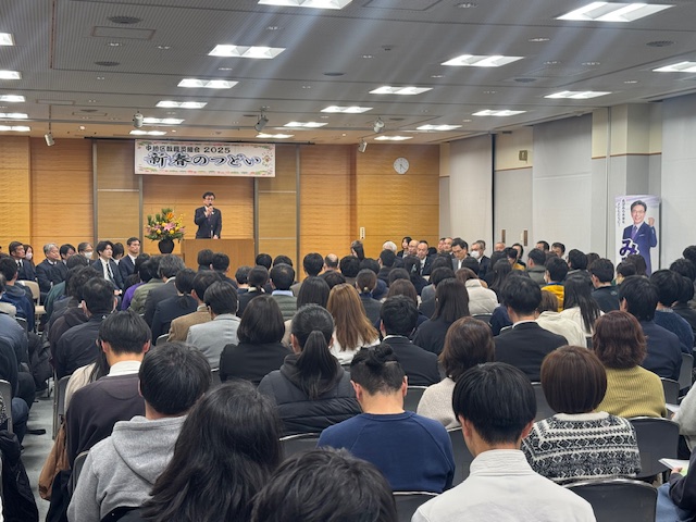 新春のつどいと書かれた横断幕が掲げられた会場で、演台に立つスーツ姿の市長が、会場に設置された椅子に座った多くの聴衆を前に挨拶をしている様子を捉えた写真