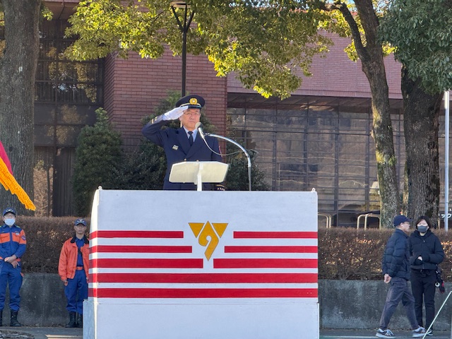 前面にオレンジ色と白の横縞に秦野市のマークが書かれた屋外の壇上で、濃紺の制服を着た男性が敬礼をしており、その後ろで青色とオレンジ色の活動服を着た隊員と観客が見ている写真