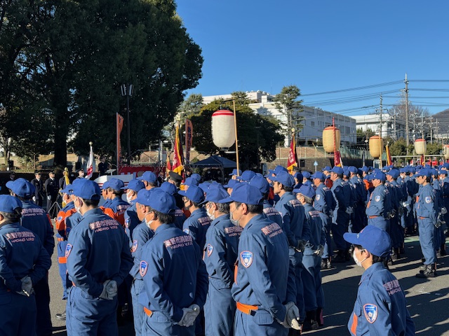 青い作業服を着た多くの消防団員が、屋外で旗や大きな提灯が飾られた広場に整列しており、式典に参加している様子を捉えた写真