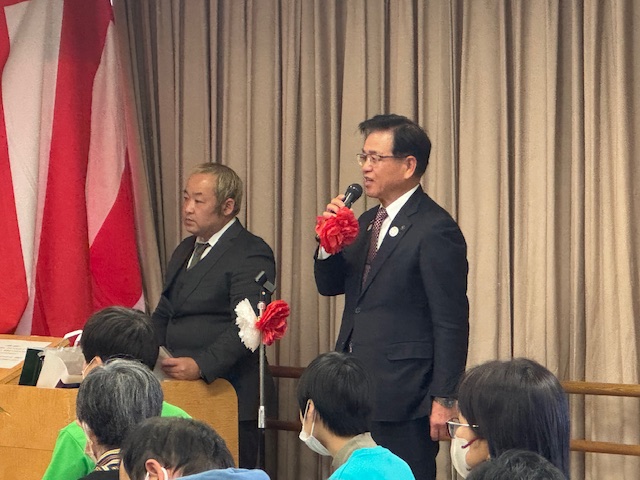 横の檀上には進行役の男性が立っており、手前に立つスーツ姿の市長が、赤い花飾りがついたマイクを手に大勢の聴衆を前に挨拶を行っている式典の様子の写真