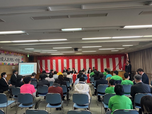 前方に紅白幕が飾られ、左側に「成人お祝いの会」と書かれ横断幕が設置された会場で、右側のマイクスタンドの前でスーツ姿の市長がマイクを持って挨拶をしており、多くの参加者が椅子に座って話を聞いている様子の写真