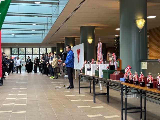 トロフィーや賞状が並んだテーブルの奥で青いジャンバー姿の市長が大勢の人の前で挨拶をしている開会式の様子を写した写真