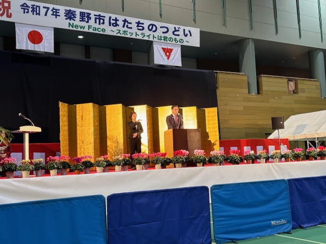 金屏風の前に設置された演台に立つスーツ姿の男性が挨拶をしている写真