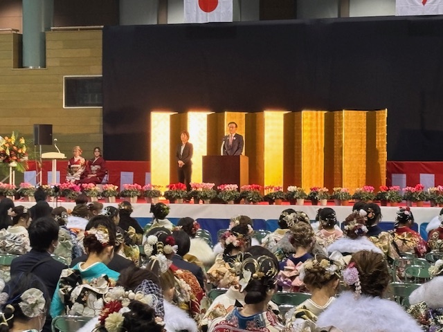 着物姿のたくさんの成人の人たちの前で金屏風の前に設置された演台に立ち挨拶をしているスーツ姿の男性の写真