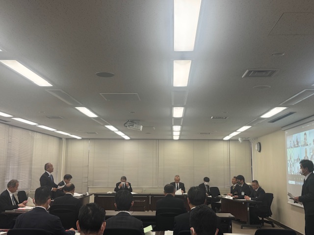 天井の蛍光灯が明るく照らす会議室で、大勢の参加者が着席し、発言者がマイクを使って話す様子を写した会議の写真