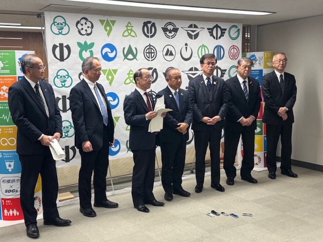 白い幕に複数のロゴが描かれた部屋で、全員がスーツを着用した7人の男性が立ち、そのうちの一人が紙を手に話している様子を捉えた写真