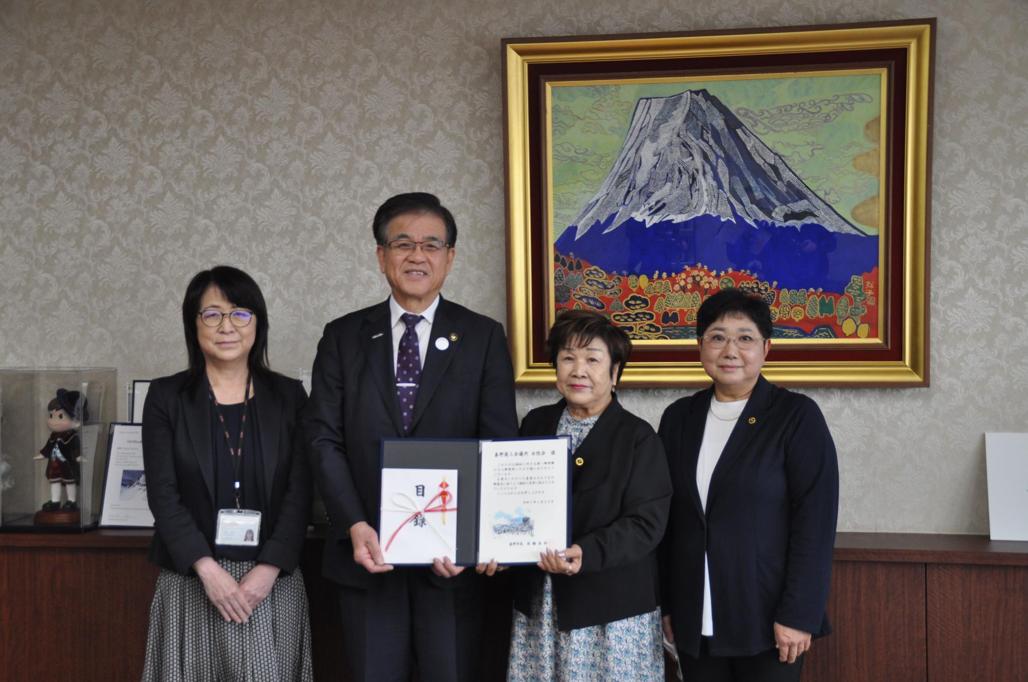 日本画風の富士山の絵が飾られた部屋で、中央の市長と隣の女性が目録などを二人で持ち、その両隣に女性二人が並んでカメラに向かって立っている記念写真