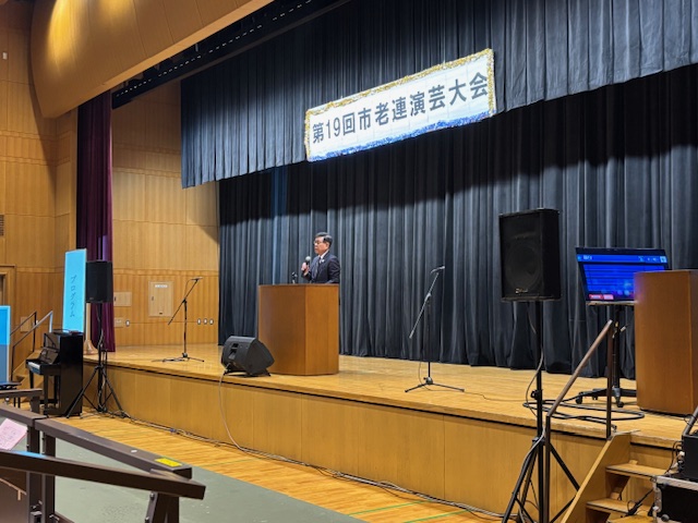 演説台に立つスーツ姿の市長が、黒い幕に「第19回市老連演芸大会」と書かれた白い横断幕を背景に話している舞台上の様子を捉えた写真