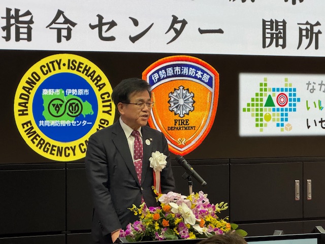 複数の市の紋章や「指令センター開所」の文字が描かれた背景の前で、マイクスタンドに立つスーツ姿の市長が、胸に白い花をつけ挨拶をしている様子を捉えた写真