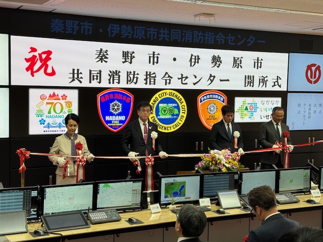 複数の市の紋章が映し出された大型モニターの前で、男女4人が赤いリボンにハサミを入れてテープカットをしている式典の様子を捉えた写真