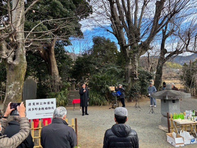 木々の間に設置された場所に立ちマイクを持って話すスーツ姿の男性を複数の観客が囲む様子を捉えた写真