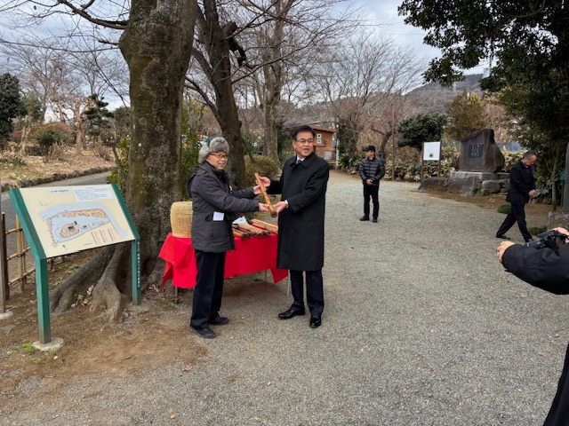 屋外の砂利道に置かれた赤い布の台の前で、茶色い尺八を持ち、お辞儀をしながら受け渡しをしている市長と男性を写した写真