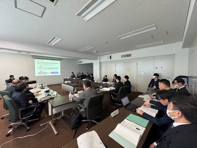 中央のモニターに資料が映し出され、コ型に設置された長机とその後方に設置された長机に座った参加者の人たちが画面を見ている審査会での写真