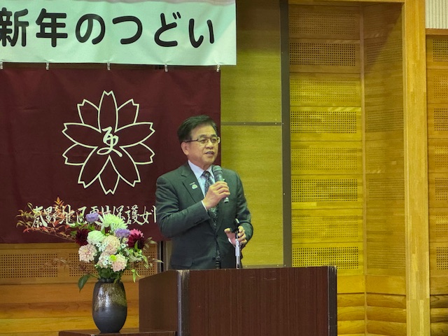 新年のつどいの幕が掲げられた壁の前で、スーツ姿の市長がきれいな花が飾られた説台に立ち、マイクを使って話している様子を写した写真