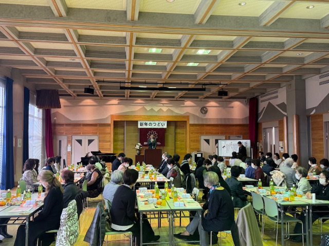 飲物や軽食が置かれた机を囲んで大勢の人が集まり、ステージ上の演説台に立つ男性の話を聞いている様子を捉えた写真