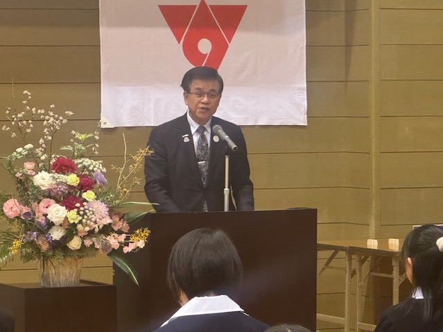 白地に赤いマークの旗が掲げられた壁の前で、スーツ姿の市長がきれいな花が飾られた説台に立ち、マイクを使って話している様子を写した写真