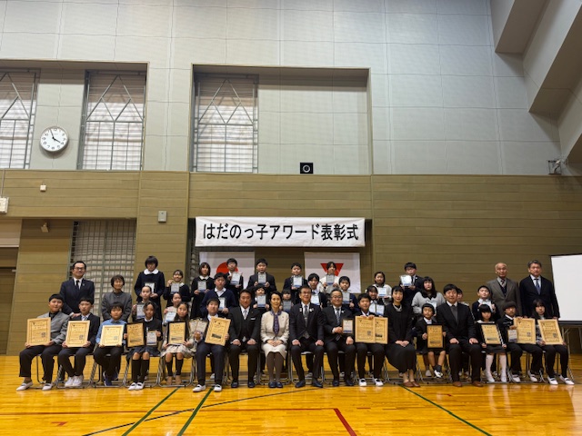 体育館のステージ上で、「はだのっ子アワード表彰式」と書かれた横断幕を背景に、賞状や盾を手にした子供たちとスーツを着た大人たちが集合した記念写真