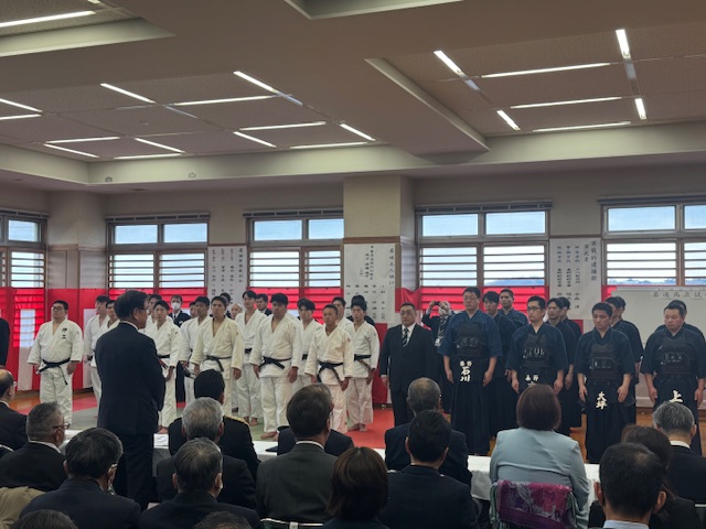 広い体育館のような場所で、柔道着や剣道着を着用した人たちが整列し、その前にいるスーツ姿の来賓たちに挨拶をしている様子を捉えた写真