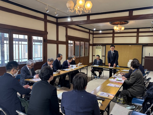 大日本報徳社の室内でロ型に設置された長机に関係者が座り、一人の男性が立って挨拶をしている写真