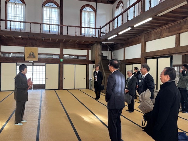 伝統的な日本家屋の畳敷きの広間で、熱心に説明をしている男性の話ををスーツ姿の複数の男性が聞いている様子を捉えた写真