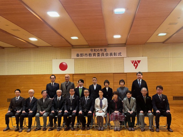 「令和6年度 秦野市教育委員会表彰式」と書かれた横断幕を背景に、スーツやワンピース姿の複数の男女が集合し1列目の椅子に11名、2列目に7名の方々が立って記念撮影をしている