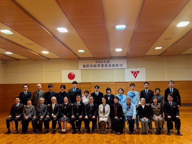 「令和6年度 秦野市教育委員会表彰式」と書かれた横断幕を背景に、スーツやワンピース姿、水色のシャツの男女が集合し1列目の椅子に13名、2列目に13名の方々が立って記念撮影をしている写真
