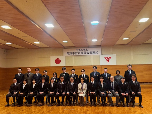 「令和6年度 秦野市教育委員会表彰式」と書かれた横断幕を背景に、スーツやワンピース姿の複数の男女が集合し1列目の椅子に13名、2列目に12名の方々が立って記念撮影をしている写真