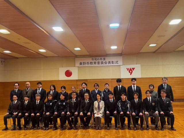 「令和6年度 秦野市教育委員会表彰式」と書かれた横断幕を背景に、スーツやワンピース姿の複数の男女が集合し1列目の椅子に13名、2列目に13名の方々が立って記念撮影をしている写真