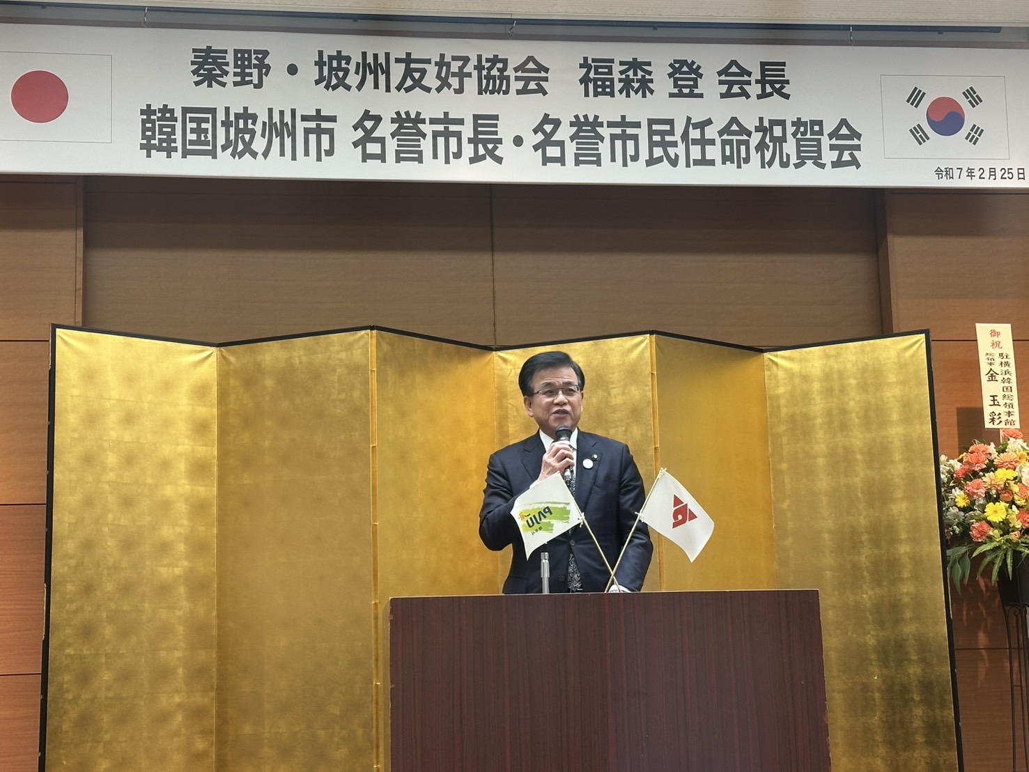 金色の屏風の前で、スーツ姿の市長が演台でマイクを手に挨拶をしている写真