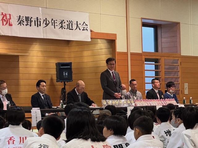 秦野市少年柔道大会で柔道着を着た少年少女たちが着席しており、前列に並ぶスーツ姿の男性のうち市長が立ち上がって挨拶をしている写真