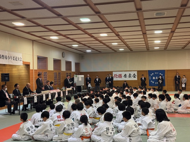 畳の上で、多くの柔道着姿の少年少女たちが座って整列しており、前方のテーブルに立つスーツ姿の男性の挨拶を聞いている様子の写真