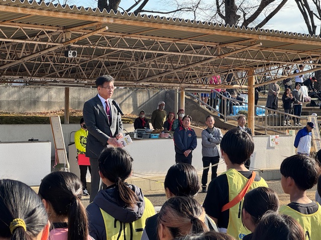 晴れた屋外の広場で、スーツ姿の市長が子供たちに向かって挨拶をしており、その背後には他の関係者や保護者らしき人々が見えるイベント風景の写真