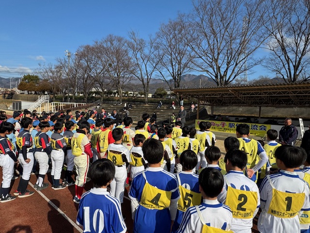 駅伝交流会で、大勢の子供たちがユニフォーム姿で整列しており、壇上に立つスーツ姿の男性がマイクを使って挨拶をしている写真