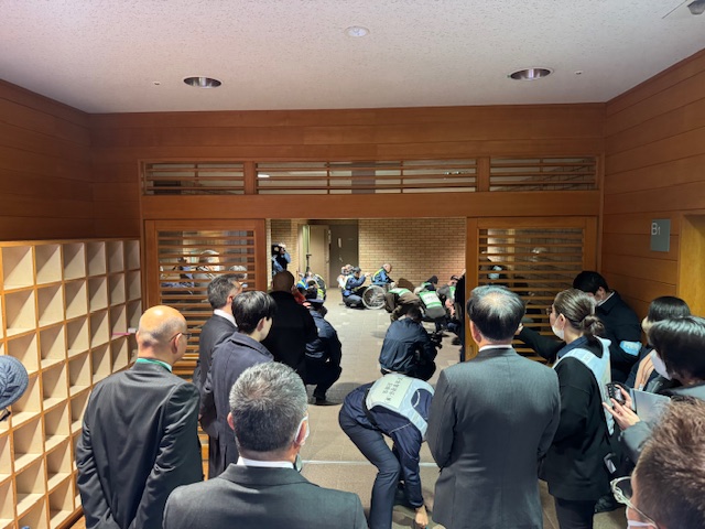 木製の仕切り越しに多くの人々が集まり、車椅子利用者と警備員らが廊下でやり取りしている様子を後方から撮影した室内の写真