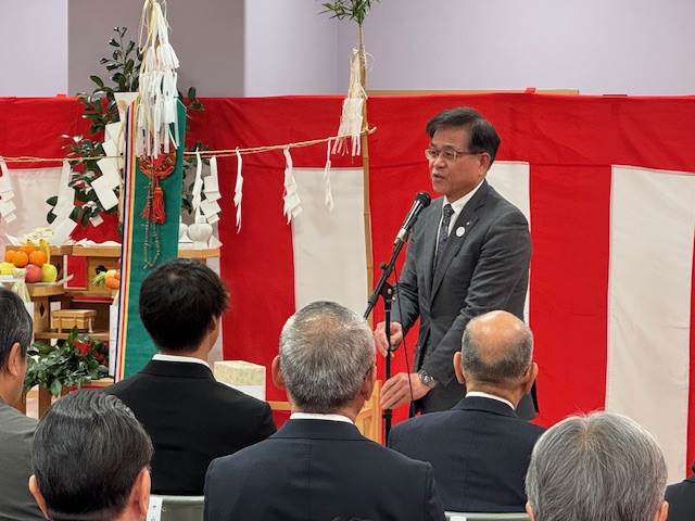 紅白の幕と神棚のような祭壇が設置された会場で、スーツ姿の市長がマイクを使って挨拶をしている様子を撮影した写真