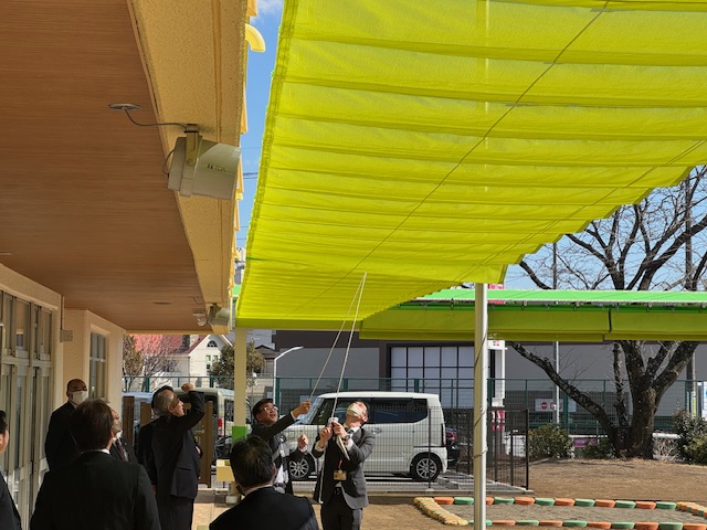 新設された黄色い日よけの開閉を、複数のスーツ姿の男性たちが屋外で試している様子を捉えた写真