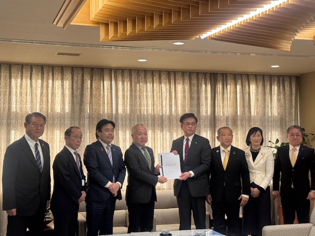 市の応接室でスーツ姿の男女8人が並んでおり、中央の市長と男性が書類を一緒に持ち、カメラに向かってポーズをとっている集合写真