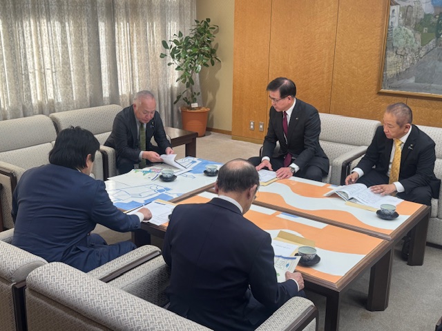 和やかな雰囲気の応接室で、5人のスーツ姿の男性が地図を広げたテーブルを囲んで座り、熱心に話し合っている様子を写した写真